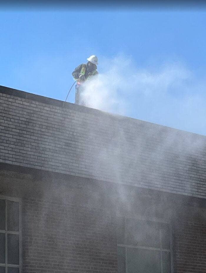 Crew member washing a brick parapet from the roof, mist drifting down the wall