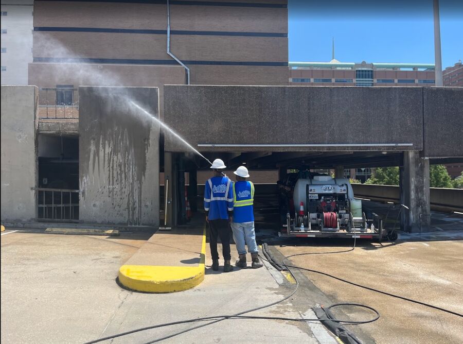 Two crew members washing black runoff stains from a parking garage wall, trailer rig parked under the deck